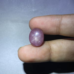 May include: A pink oval-shaped gemstone with a star-like pattern inside. The gemstone is held by a finger.