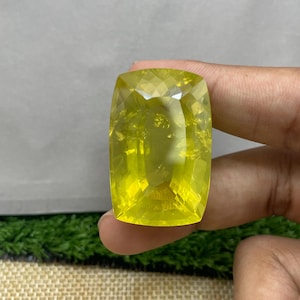 Może przedstawiać: Duży, fasetowany, prostokątny kamień szlachetny z zaokrąglonymi rogami. Klejnot ma żywy żółty kolor i wydaje się być trzymany przez osobę. Powierzchnia klejnotu odbija światło, ukazując jego wewnętrzną strukturę i przejrzystość.