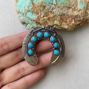 May include: A silver-toned horseshoe pendant with eight round, bright turquoise stones set in a curved design. The pendant has a detailed, textured surface and is likely a piece of jewelry.