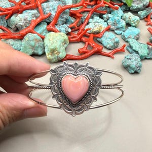 May include: A silver cuff bracelet with a pink heart-shaped stone set in a detailed frame. The bracelet has a double-band design. The background includes red coral and turquoise stones. The bracelet is approximately 15 cm in diameter.