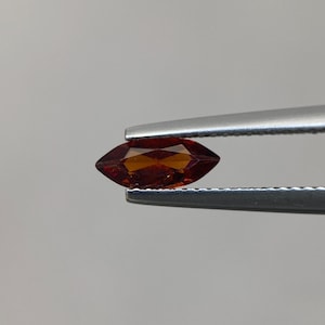 Puede incluir: Una gema facetada en forma de marquesa, sujeta con pinzas. La gema es de color marrón rojizo oscuro con una superficie pulida. La forma es alargada con extremos puntiagudos, y las facetas captan la luz, creando un efecto brillante.