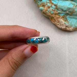 Op de afbeelding: Een zilveren ring met een turkoois inlegwerk en geometrisch ontwerp. De ring heeft een band van turkooizen stenen en zilveren accenten. De ring wordt in een hand gehouden, met een stuk turkoois steen op de achtergrond.