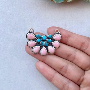 May include: A silver pendant featuring a fan-shaped arrangement of teardrop-shaped pink stones and smaller round turquoise stones. The metal setting has a decorative rope-like border and two small circular loops at the top for attachment.