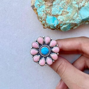 May include: A silver ring featuring a central bright blue circular stone surrounded by a ring of twelve teardrop-shaped light pink stones. The metal setting has a twisted rope texture around the center stone and a polished silver finish.