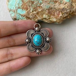 May include: A silver pendant with a turquoise stone center. The pendant has a floral design with a rope border around the stone and radiating lines on the petals. The pendant is held in a hand.