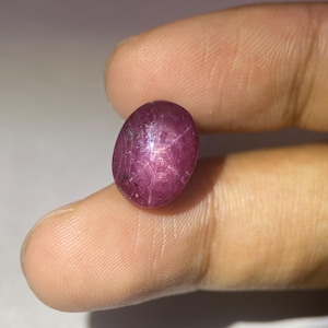 Könnte beinhalten: Ein ovaler Sternrubin-Edelstein. Der Edelstein hat eine tiefrote bis violette Farbe und zeigt einen sechsstrahligen Stern auf seiner Oberfläche. Der Stein wird zwischen den Fingern gehalten, was seine glatte Textur und sein natürliches Aussehen hervorhebt.