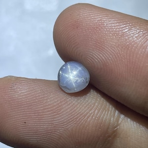 May include: A small, round, pale blue gemstone with a six-rayed star pattern. The gem is held between two fingers, showcasing its polished surface and the star effect created by light refraction. The stone has a smooth, rounded shape.