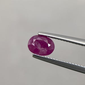 Könnte beinhalten: Ein facettierter, ovalförmiger Rubin, gehalten von einer silbernen Pinzette. Der Rubin zeigt eine tiefe, gesättigte rote Farbe mit mehreren Facetten, die das Licht einfangen. Der Hintergrund ist ein schlichtes, neutrales Grau.