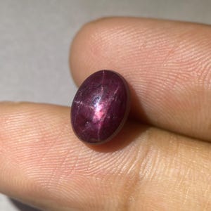 May include: A polished, oval-shaped gemstone with a deep burgundy color and a star-like effect. The gem has a smooth surface and a rich, saturated hue. The star effect is visible in the center of the stone, creating a captivating visual.