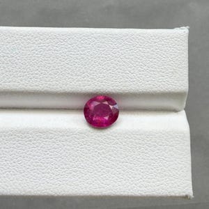 May include: A round, faceted ruby gemstone, displaying a vibrant red hue. The gemstone is set against a white, textured background. The ruby's polished surface reflects light, showcasing its clarity and brilliance. A close-up view highlights the gem's facets.