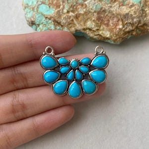 May include: A silver pendant featuring a cluster of bright turquoise teardrop-shaped stones. The stones are set in a silver metal frame, creating a striking contrast. The pendant has two small loops at the top for attaching to a chain.