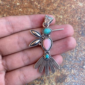 May include: A silver hummingbird pendant with turquoise and pink gemstones. The pendant features a detailed design with a teardrop-shaped pink stone in the centre, flanked by turquoise stones and silver wings and tail.