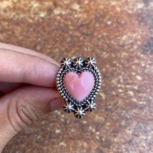 Könnte beinhalten: Ein silberner Ring mit einem rosa herzförmigen Stein. Der Ring hat ein detailliertes Design mit einem geflochtenen Rand um das Herz und sternförmigen Akzenten. Der Ring wird von einer Hand gehalten.