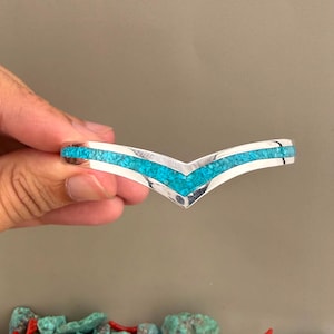 May include: A V-shaped cuff bracelet made of polished silver metal featuring a continuous inlay of small, crushed bright turquoise blue stones that follow the chevron design along the centre of the band, creating a smooth and reflective surface.