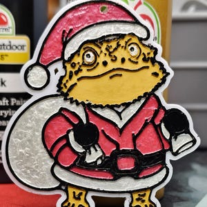 Puede incluir: Adorno navideño con un dragón barbudo vestido de Papá Noel. El adorno tiene un gorro y un traje de Papá Noel rojos con ribetes blancos, un cinturón negro y un saco blanco. El lagarto es amarillo y tiene ojos negros.