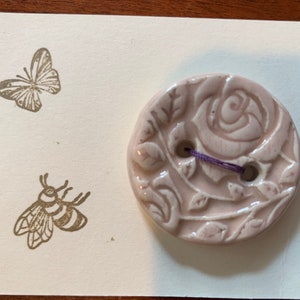 May include: A pale pink ceramic button with a raised design of a rose and leaves. The button has two holes and a purple thread running through them.