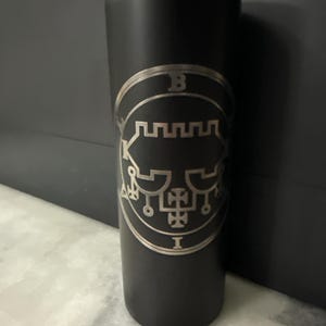 Puede incluir: Un vaso de acero inoxidable negro con un sello plateado del demonio Bune grabado en el lateral. El sello está rodeado de un círculo con las letras "B" e "I" en la parte superior e inferior.