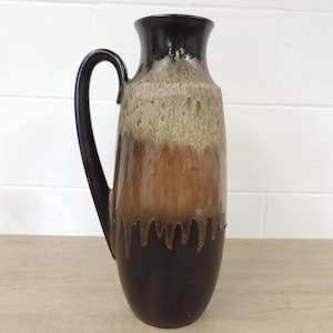 May include: A tall ceramic vase with a dark brown handle and a gradient colour scheme. The top is black, transitioning to beige, then to a rich brown, and finally to a dark brown base. The vase has a drip glaze effect.