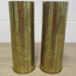 May include: A pair of brass vases with etched floral patterns. The cylindrical vases have a scalloped top and a flared base. The etched design includes large leaves and flowers. They are displayed on a light-coloured wooden surface.