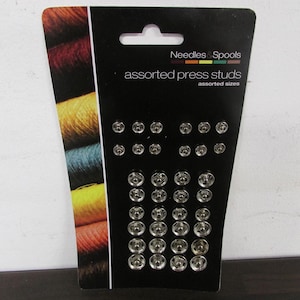 May include: A black card displaying assorted press studs in various sizes. The card is labelled "Needles & Spools" and "assorted press studs". The left side of the card features a colourful display of threads.