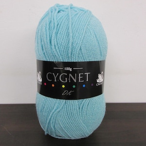 May include: A ball of light blue Cygnet DK yarn. The black label displays the brand name and a swan logo. The label also indicates the yarn weight is 100g. The yarn is tightly wound, showing the texture of the fibres.