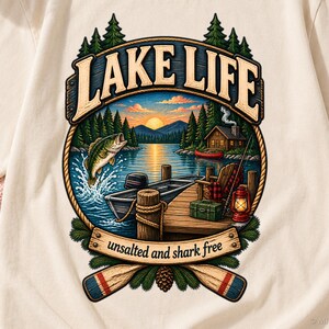 May include: A cream-coloured t-shirt featuring a graphic of a lake scene with a jumping fish, a boat at a wooden dock, a cabin, and a lantern. The text reads LAKE LIFE and unsalted and shark free, framed by pine trees and two wooden oars.