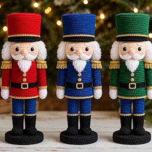 Puede incluir: Tres muñecos cascanueces de ganchillo alineados. Cada muñeco presenta un sombrero alto, una barba blanca y un uniforme con ribetes dorados. El muñeco de la izquierda es rojo, el del centro es azul y el de la derecha es verde.