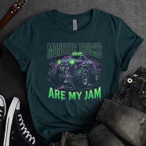 Pu&ograve; includere: T-shirt verde scuro con grafica di monster truck verde neon e viola e la scritta "MONSTER TRUCKS ARE MY JAM". La grafica include un'illustrazione dettagliata di un monster truck con pneumatici grandi e un volto ringhioso.