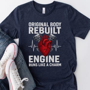 May include: Navy blue t-shirt with the text "ORIGINAL BODY REBUILT ENGINE RUNS LIKE A CHARM". The graphic features a red heart, pistons, and a heartbeat line. White sneakers and blue jeans are visible.