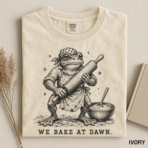 Könnte beinhalten: Ein elfenbeinfarbenes T-Shirt mit einer schwarzen Tuschezeichnung eines Krötenwesens mit Bandana und Schürze, das ein Nudelholz neben einer Rührschüssel hält. Der Text "WE BAKE AT DAWN." steht unter dem Motiv. Das Shirt hat ein Comfort Colors Etikett.