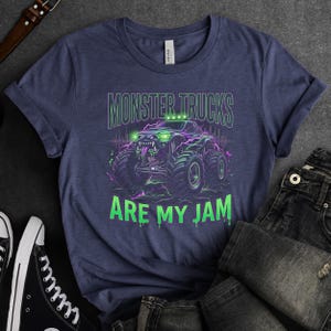 Pu&ograve; includere: T-shirt blu navy con grafica di monster truck e la scritta "MONSTER TRUCKS ARE MY JAM" in verde e viola. Il design presenta un monster truck con ruote grandi e un'espressione minacciosa.