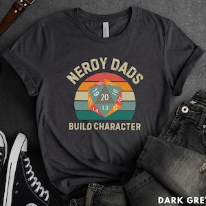 May include: Dark grey t-shirt with a vintage-style graphic of a twenty-sided die and the text "Nerdy Dads Build Character".