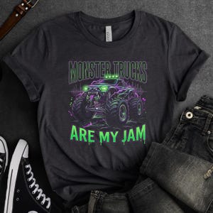 Pu&ograve; includere: T-shirt grigio scuro con una grafica di un monster truck e la scritta "MONSTER TRUCKS ARE MY JAM". Il monster truck &egrave; raffigurato in verde e viola con ruote grandi e un design minaccioso.