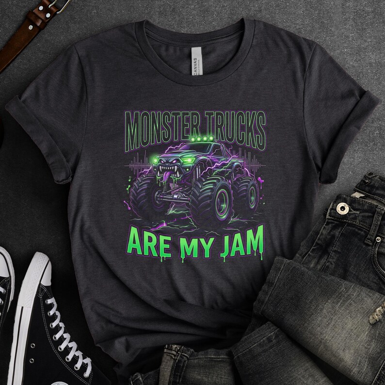 Pu&ograve; includere: T-shirt grigio scuro con una grafica di un monster truck e la scritta "MONSTER TRUCKS ARE MY JAM". Il monster truck &egrave; raffigurato in verde e viola con ruote grandi e un design minaccioso.