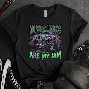 Pu&ograve; includere: T-shirt nera con grafica di monster truck verde e viola, con la scritta "MONSTER TRUCKS ARE MY JAM". Il monster truck ha pneumatici grandi e un design minaccioso.