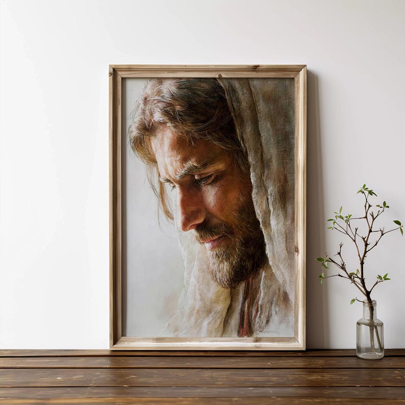 May include: A framed portrait of a man with long brown hair and a beard, wearing a white cloth draped over his head. The artwork is in a wooden frame and is displayed on a wooden surface.