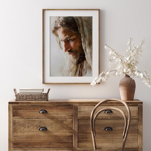 May include: A framed portrait of a man with long hair and a beard, wearing a draped cloth. The artwork is displayed on a wooden dresser with a vase of white flowers. The room has a neutral color palette.
