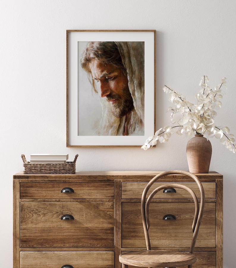 May include: A framed portrait of a man with long hair and a beard, wearing a draped cloth. The artwork is displayed on a wooden dresser with a vase of white flowers. The room has a neutral color palette.