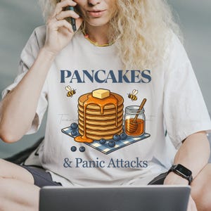 Könnte beinhalten: Weißes T-Shirt mit dem Text "PANCAKES & Panic Attacks" und einer Grafik von Pfannkuchen mit Butter, Honig, Blaubeeren und Bienen. Das T-Shirt wird von einer Person getragen.