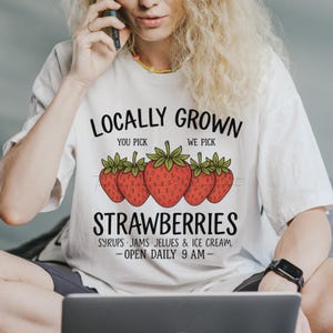 May include: White t-shirt with the text "LOCALLY GROWN STRAWBERRIES" and a graphic of five red strawberries. The shirt also lists "SYRUPS JAMS JELUES & ICE CREAM" and "OPEN DAILY 9 AM".