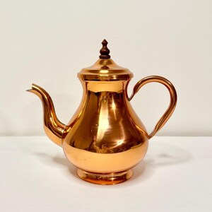 May include: A copper teapot with a wooden knob on the lid. The teapot has a rounded body and a spout on the side.