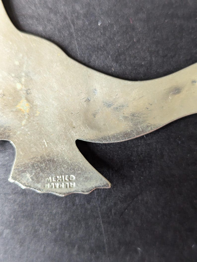 Może przedstawiać: Srebrny metalowy wisiorek w kształcie ptaka. Wisiorek ma gładką powierzchnię z pewnymi śladami użytkowania. Słowa "MEXICO" i "ALPACCA" są wytłoczone na spodzie wisiorka. Wisiorek znajduje się na ciemnym tle.