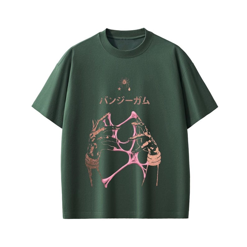 Pu&ograve; includere: T-shirt verde scuro con girocollo. La parte anteriore presenta un design grafico di mani che tengono una sostanza rosa e appiccicosa, con testo giapponese sopra. Un disegno a stella e goccia &egrave; sopra il testo.