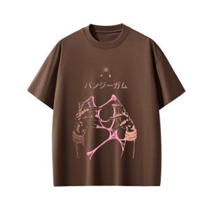 Pu&ograve; includere: T-shirt marrone con girocollo e maniche corte. La parte anteriore presenta una grafica rosa di mani che tengono una sostanza rosa gocciolante, con testo giapponese e un piccolo disegno a stella sopra. Una t-shirt unisex casual.