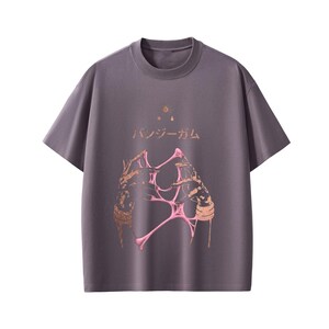 Pu&ograve; includere: T-shirt grigio a maniche corte e girocollo. La parte anteriore presenta un design grafico di mani con elementi rosa e marroni e testo giapponese. Il design include stelle e una goccia.