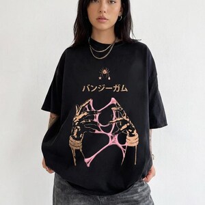 Pu&ograve; includere: T-shirt nero con un design grafico con testo giapponese e un'illustrazione rosa e beige di mani che interagiscono con una sostanza rosa e appiccicosa. La maglietta ha un girocollo e maniche corte.