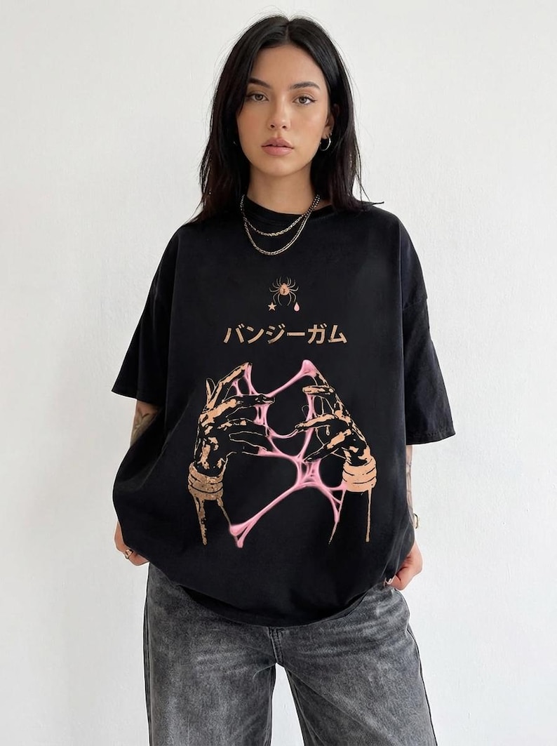 Pu&ograve; includere: T-shirt nero con un design grafico con testo giapponese e un'illustrazione rosa e beige di mani che interagiscono con una sostanza rosa e appiccicosa. La maglietta ha un girocollo e maniche corte.