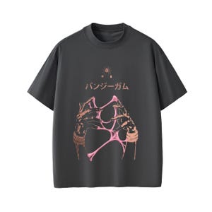 Pu&ograve; includere: T-shirt grigio scuro con girocollo. La parte anteriore presenta una grafica di mani che tengono materiale rosa e appiccicoso, con testo giapponese e un piccolo disegno di stella e ragno sopra. Le maniche sono corte.