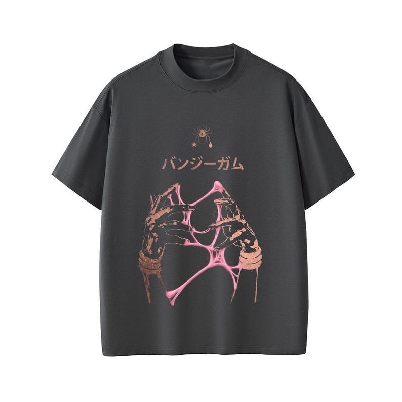 Pu&ograve; includere: T-shirt grigio scuro con girocollo. La parte anteriore presenta una grafica di mani che tengono materiale rosa e appiccicoso, con testo giapponese e un piccolo disegno di stella e ragno sopra. Le maniche sono corte.