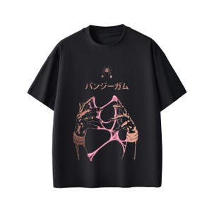 Pu&ograve; includere: T-shirt nera con un design grafico che presenta mani e materiale rosa appiccicoso. Il testo giapponese "バンジーガム" &egrave; sopra le mani e una piccola grafica di ragno &egrave; nella parte superiore.